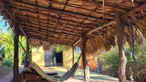 Un espacio al aire libre con techo de paja, una hamaca, palmeras y un sendero que lleva a un edificio.