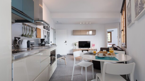 A modern kitchen with white cabinets, a dining area with a table and chairs, and a television. - Loft Gran Canaria (Arucas)