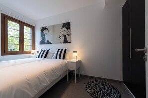 Um quarto com cama, uma mesa de cabeceira com luminária, uma obra de arte em preto e branco na parede, uma janela com persianas e uma porta.