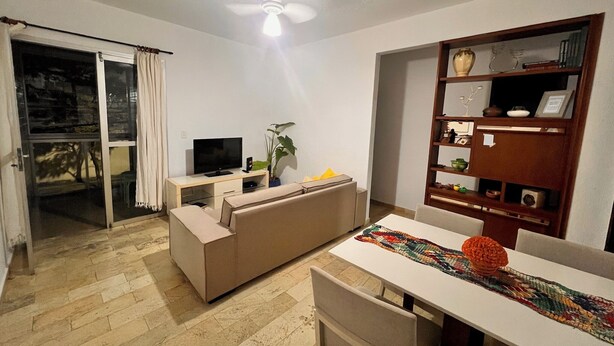 Una sala de estar con sofá, televisor y estantería. Hay una mesa de comedor con sillas y un ventilador de techo.