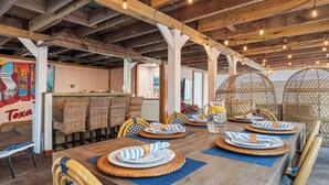 Un coin repas avec une longue table en bois, dressée pour quatre personnes, entourée de chaises en osier et agrémenté d’un plafond rustique avec des poutres apparentes et des luminaires suspendus.