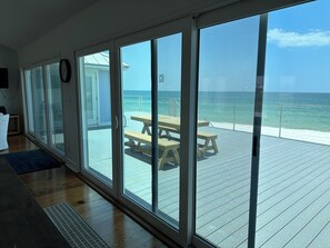Habitación con grandes puertas de vidrio corredizas que dan a una terraza exterior con mesa y bancos de madera, con vista al océano.