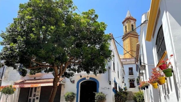 A narrow street with a tree in the foreground, a building with a blue archway, and a tower with a clock. - Casa SoHo y Alex, Apartment 3B- 2 bedrooms (Estepona)