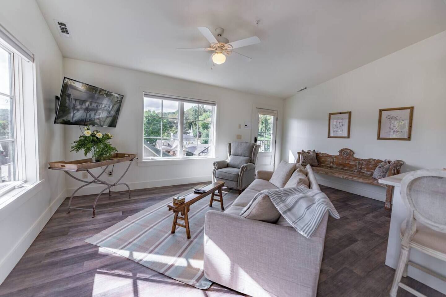 A spacious living room with a grey sofa, a wooden coffee table, and a TV mounted on the wall. There are large windows allowing natural light and a ceiling fan.