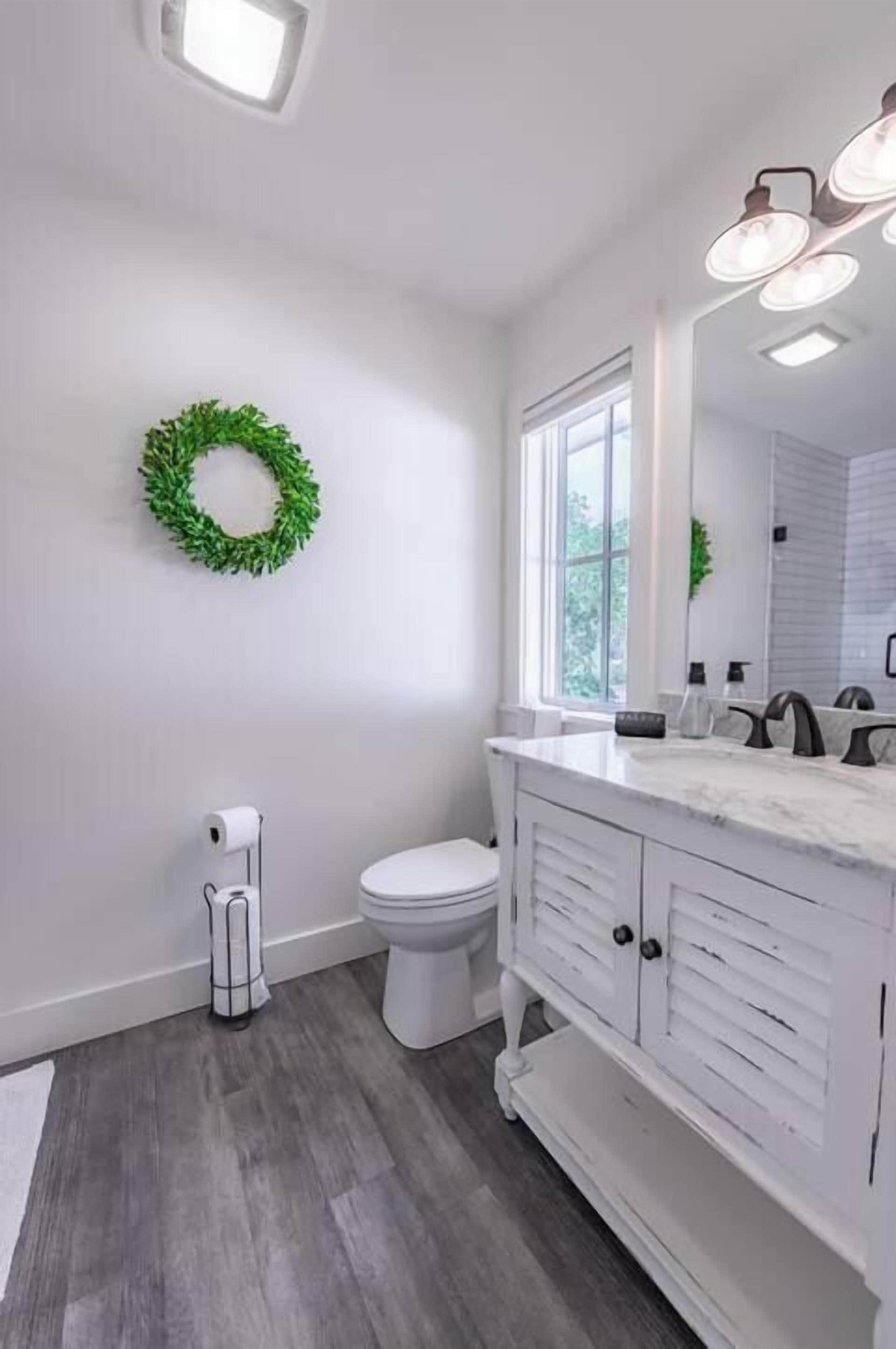 Ein Badezimmer mit weißem Waschtisch, Toilette, einem grünen Kranz an der Wand und einem Fenster, das natürliches Licht hereinlässt.