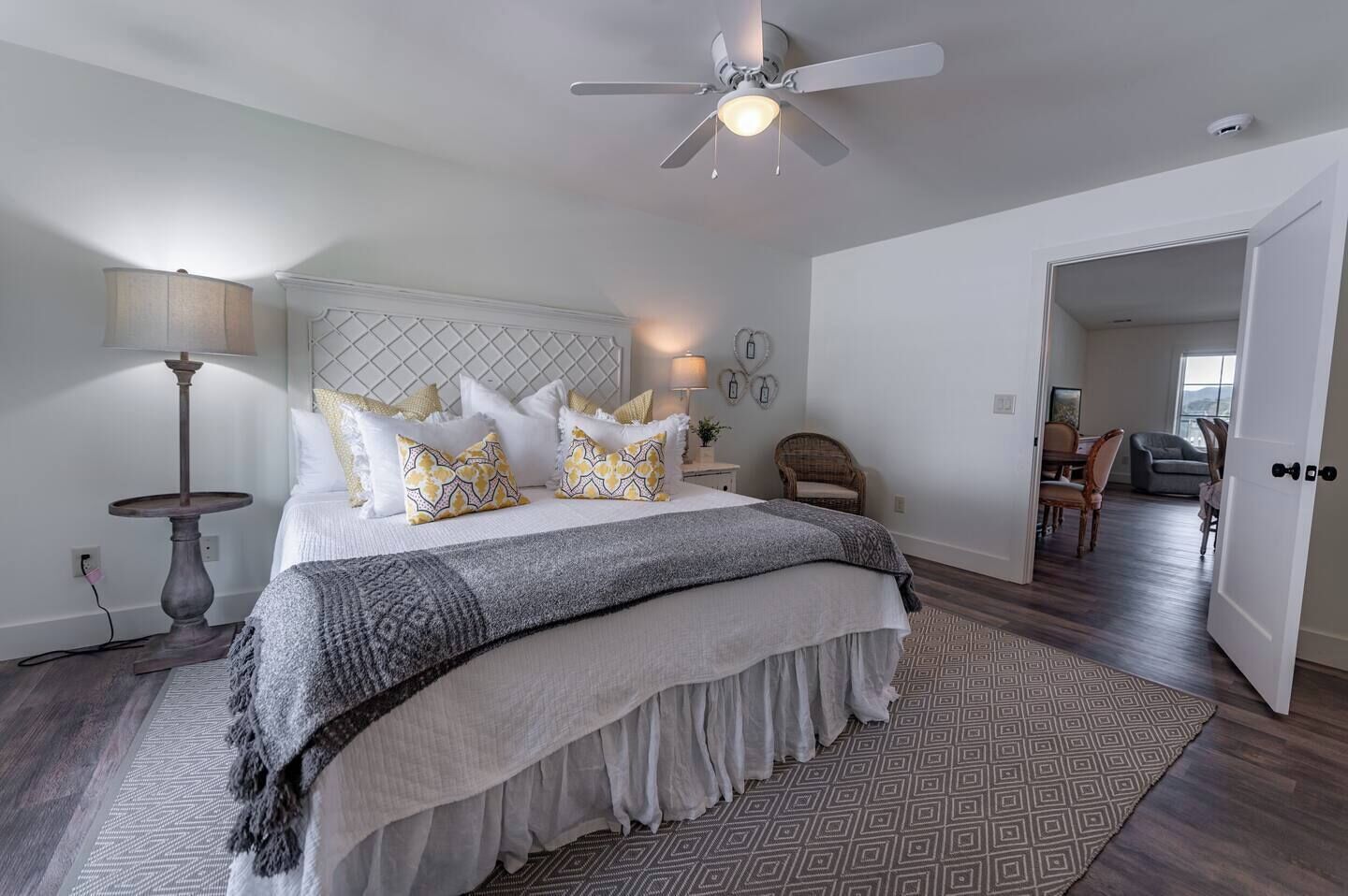 A bedroom with a bed, a ceiling fan, a nightstand, a lamp, a chair, and a view of a dining area.