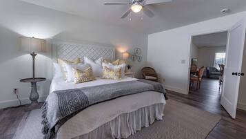 A bedroom with a bed, a ceiling fan, a nightstand, a lamp, a chair, and a view of a dining area.
