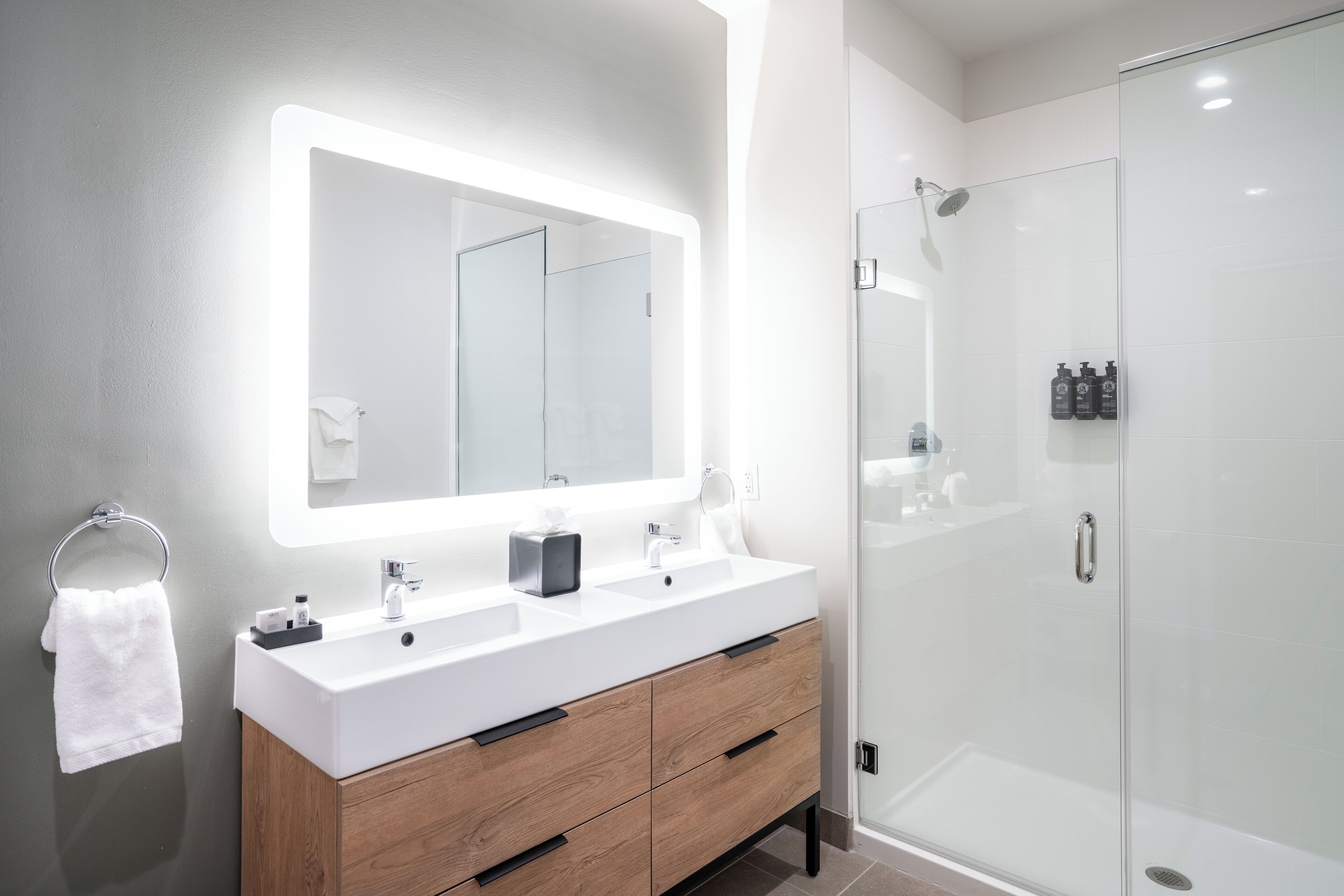 Un baño moderno con un espejo grande, dos lavamanos, un mueble de tocador con cajones y una cabina de ducha de vidrio.