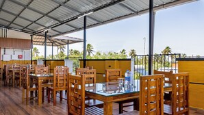 A covered outdoor dining area with wooden tables and chairs, large windows, and a view of palm trees. - Treebo Vsaffron Regency, Punnamada Lake (Ambalapuzha)