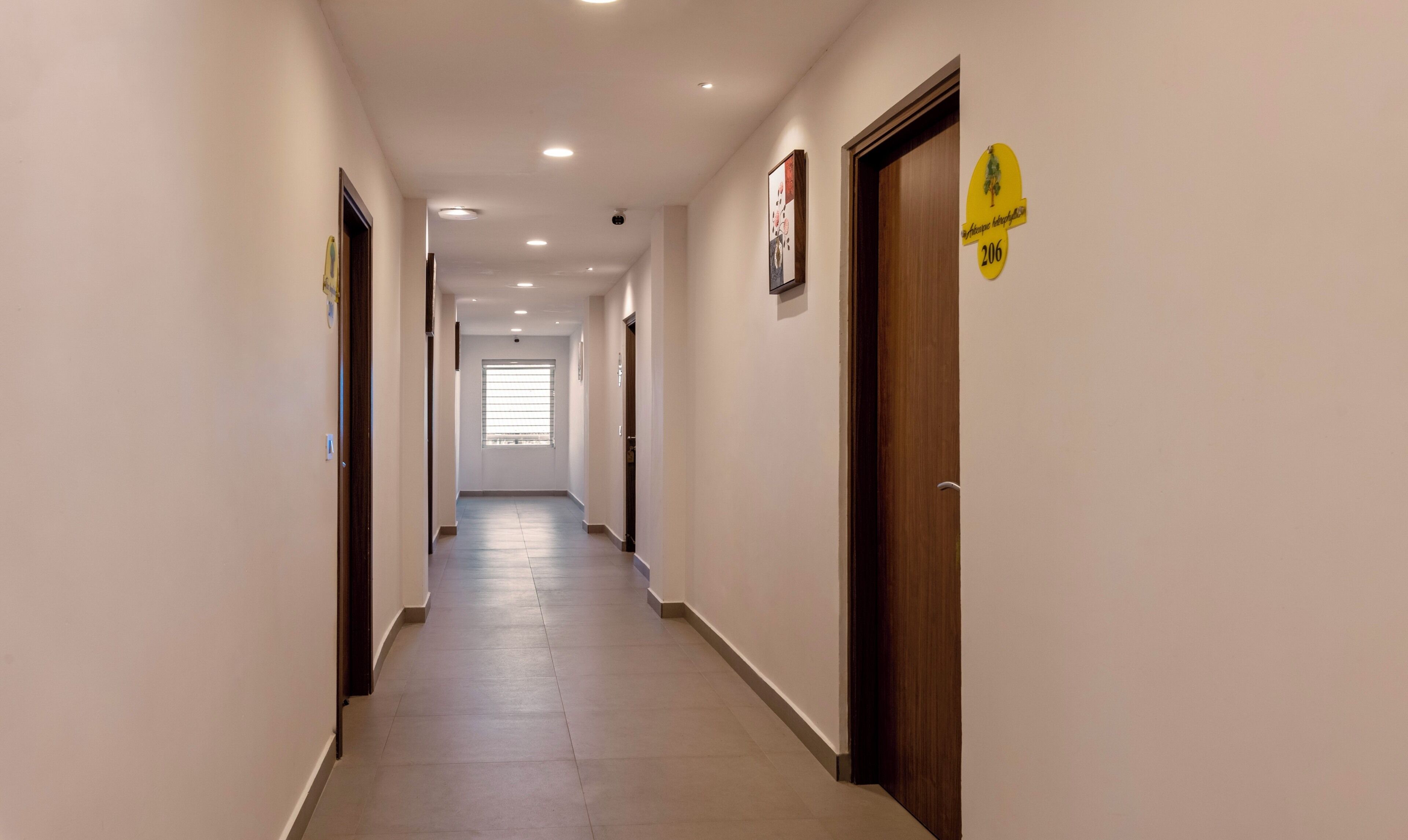 A long, narrow hallway with doors on both sides, a window at the end, and a yellow sign on the right wall.