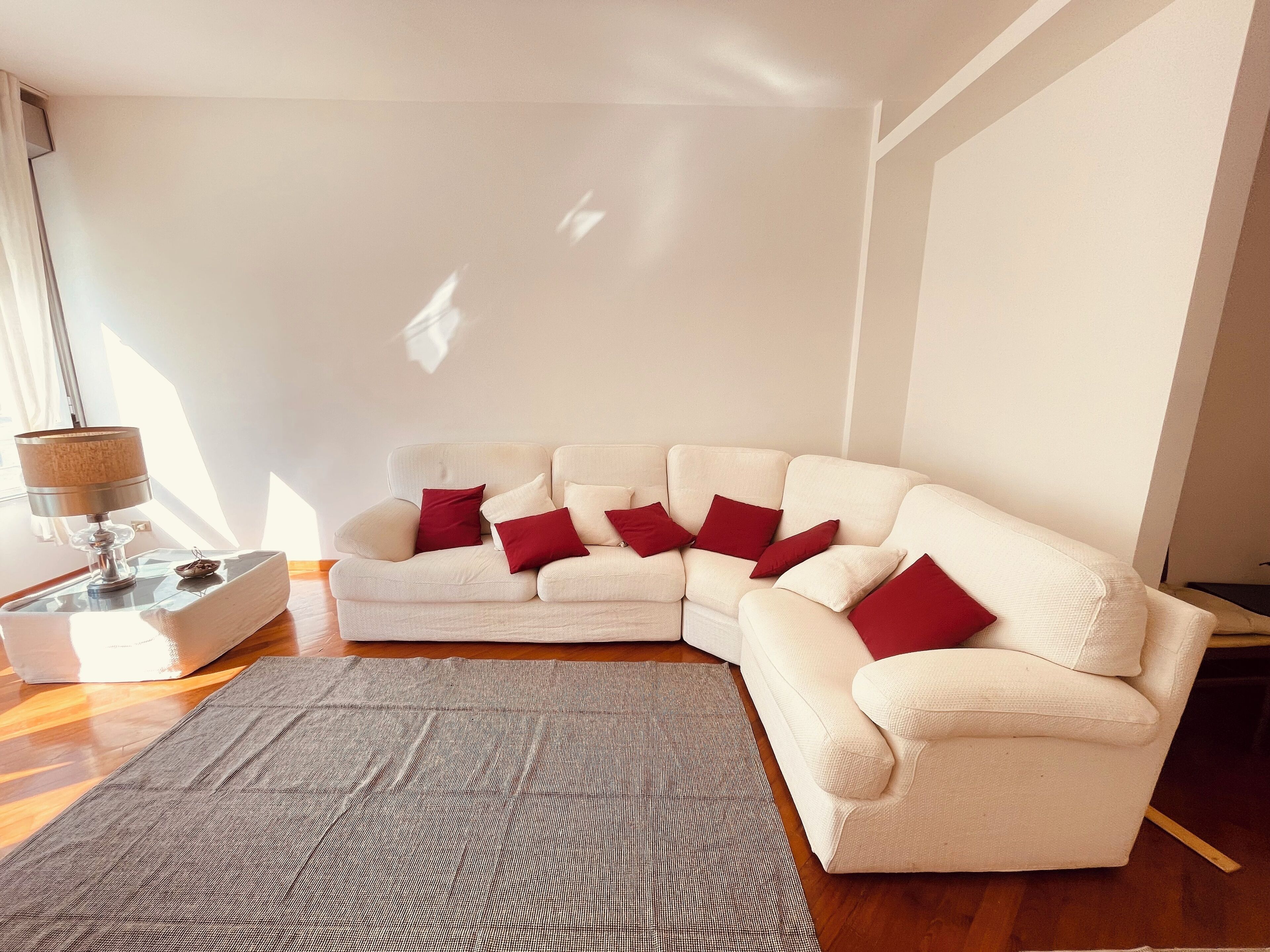Un salon avec un canapé d’angle blanc, un tapis gris et une lampe sur une table d’appoint.