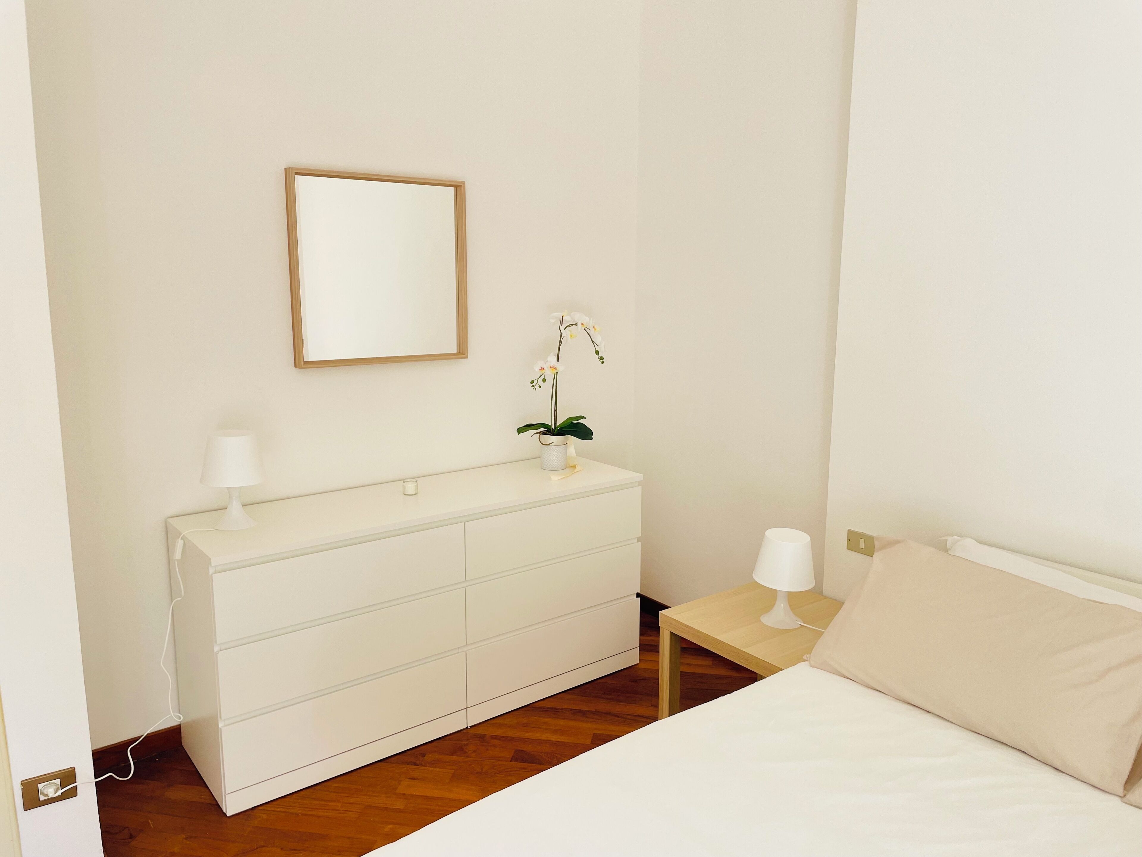 Un dormitorio con una cama, un tocador, una mesita de noche, una lámpara, un espejo y una orquídea en maceta.
