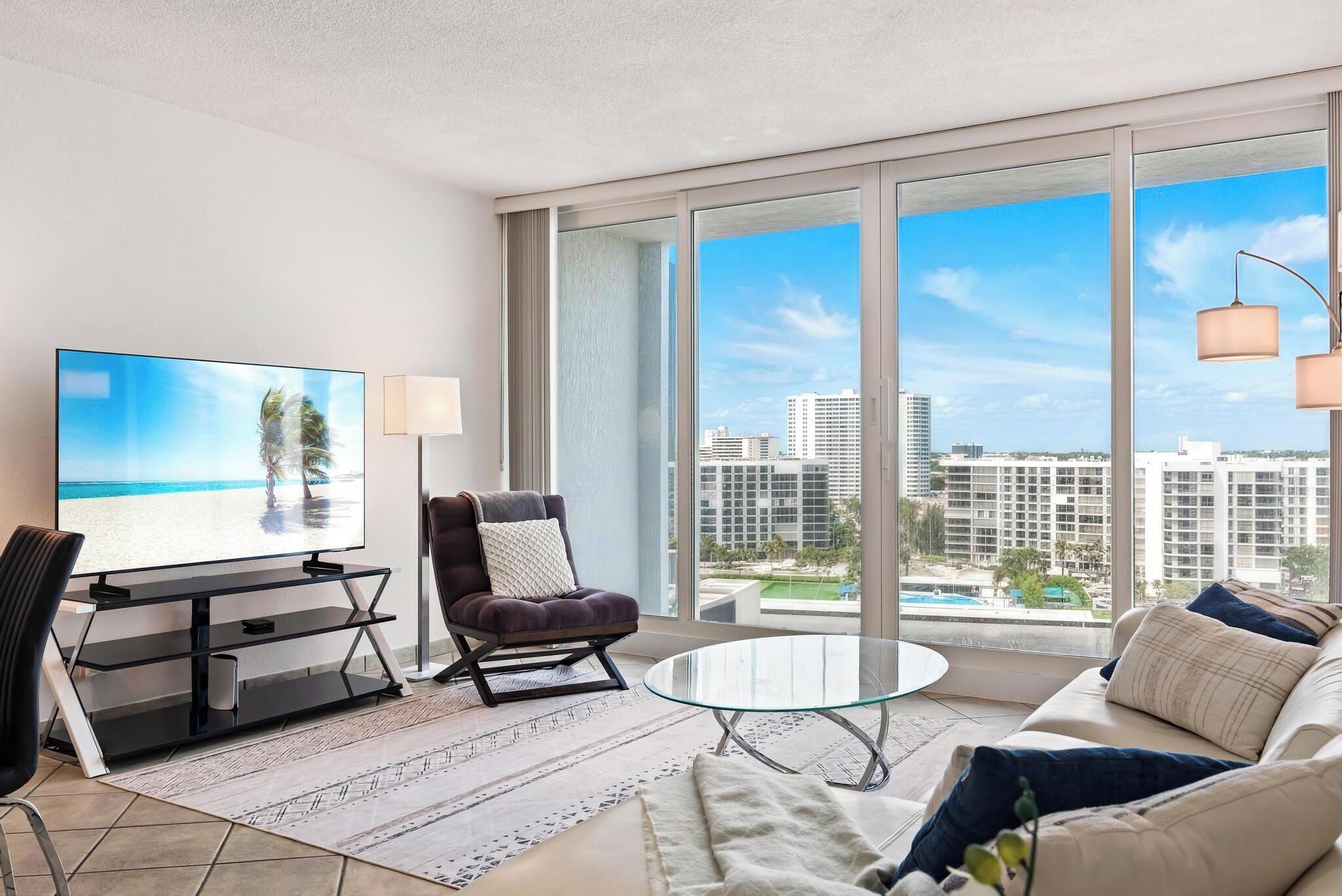 A modern living room with a large TV, comfortable seating, a glass coffee table, and a view of a cityscape through large windows.