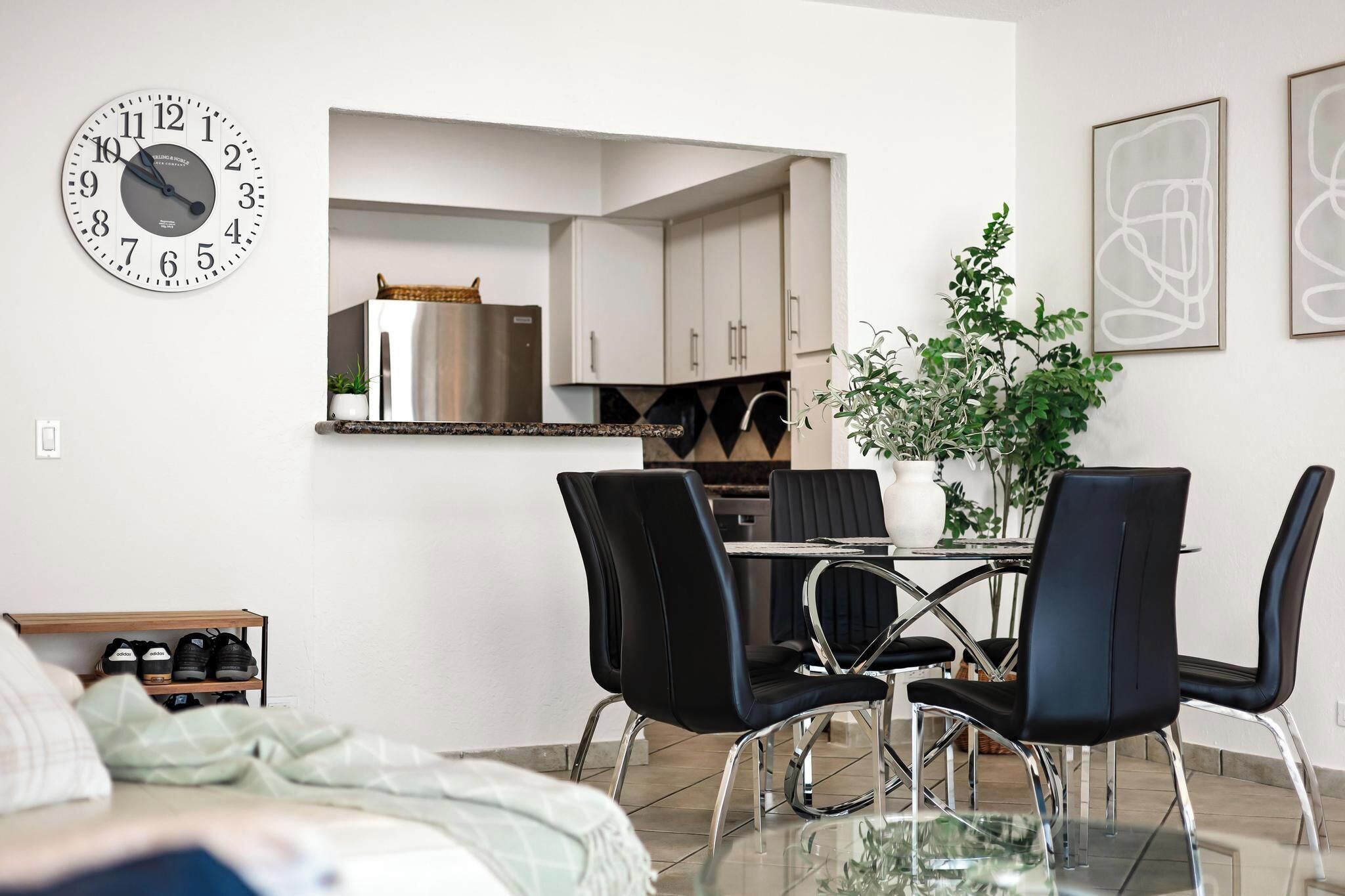 A modern living room with a glass coffee table, black chairs, a clock, and a view into the kitchen.
