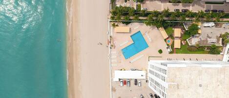 Vista aérea de um hotel à beira-mar com piscina, área de praia e estacionamento.