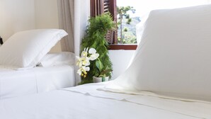 Hotellihuone, jossa on kaksi valkoista sänkyä, ruukkukasvi ja ikkuna, josta avautuu näkymä vehreään maisemaan.
