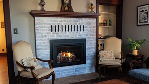 Ein Wohnzimmer mit Kamin aus Backstein, zwei Polsterstühlen, einem Holztisch und einem Bücherregal mit Büchern und Dekoration.