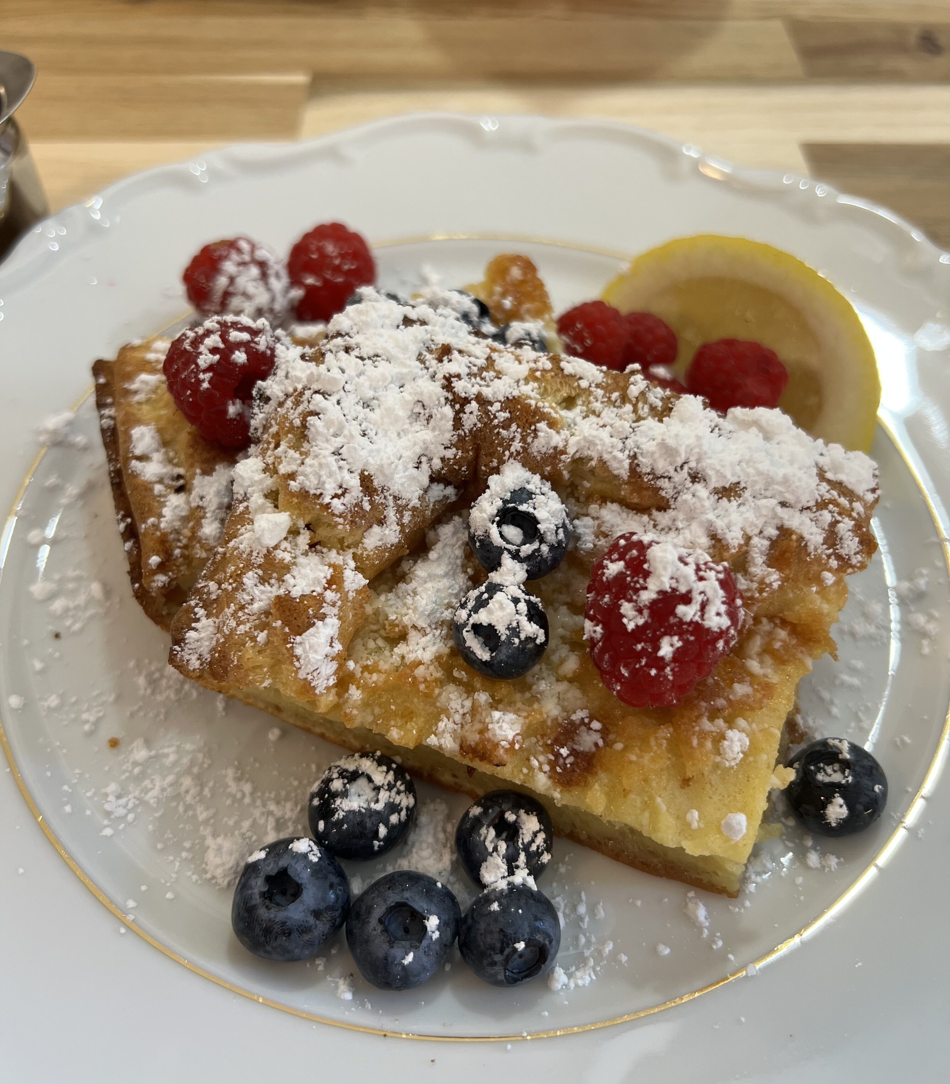 A plate of French toast with blueberries and raspberries, dusted with powdered sugar, on a white plate with a gold rim.