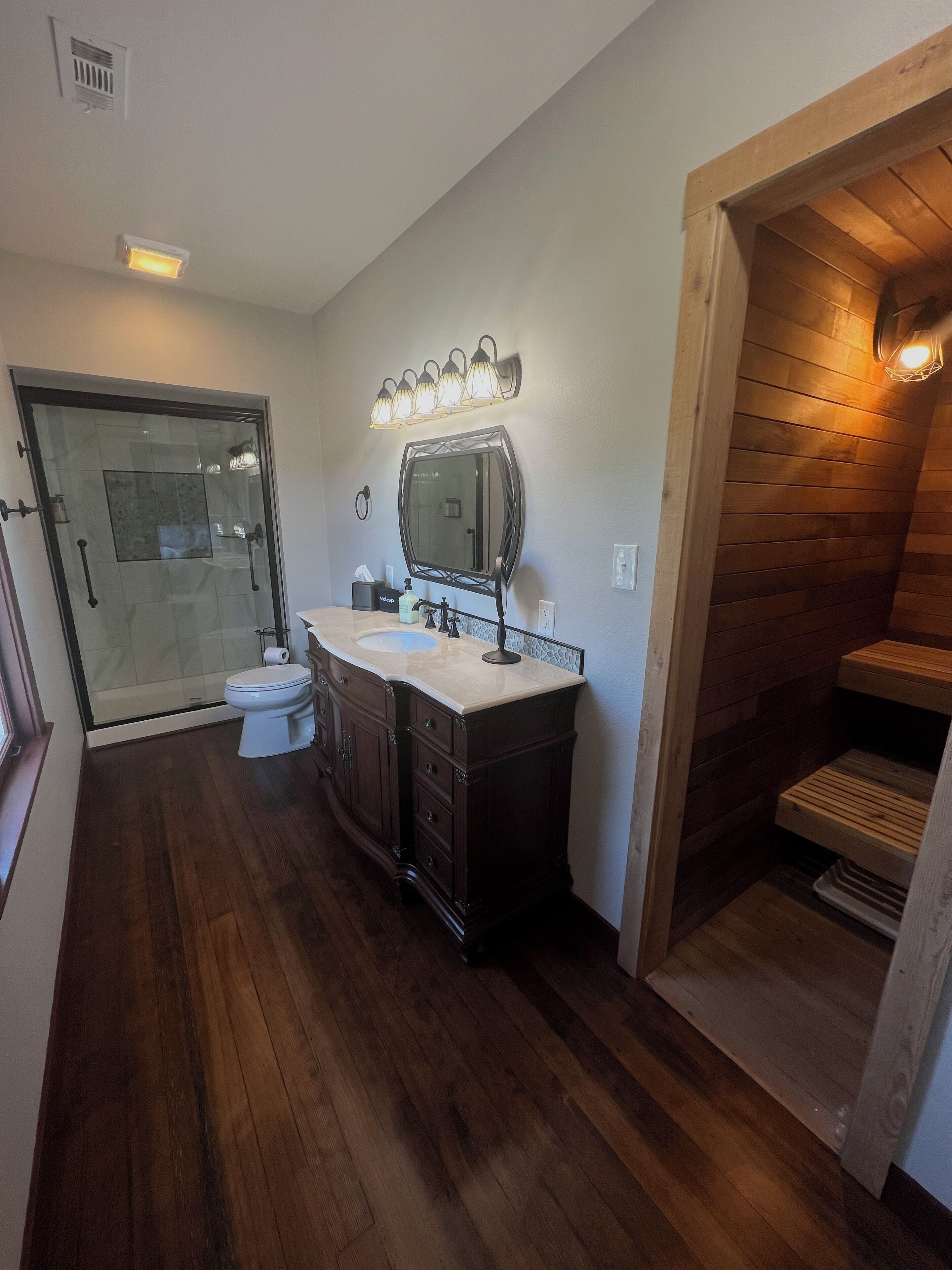 A bathroom with a glass shower enclosure, a wooden vanity with a mirror, and a wooden sauna area.