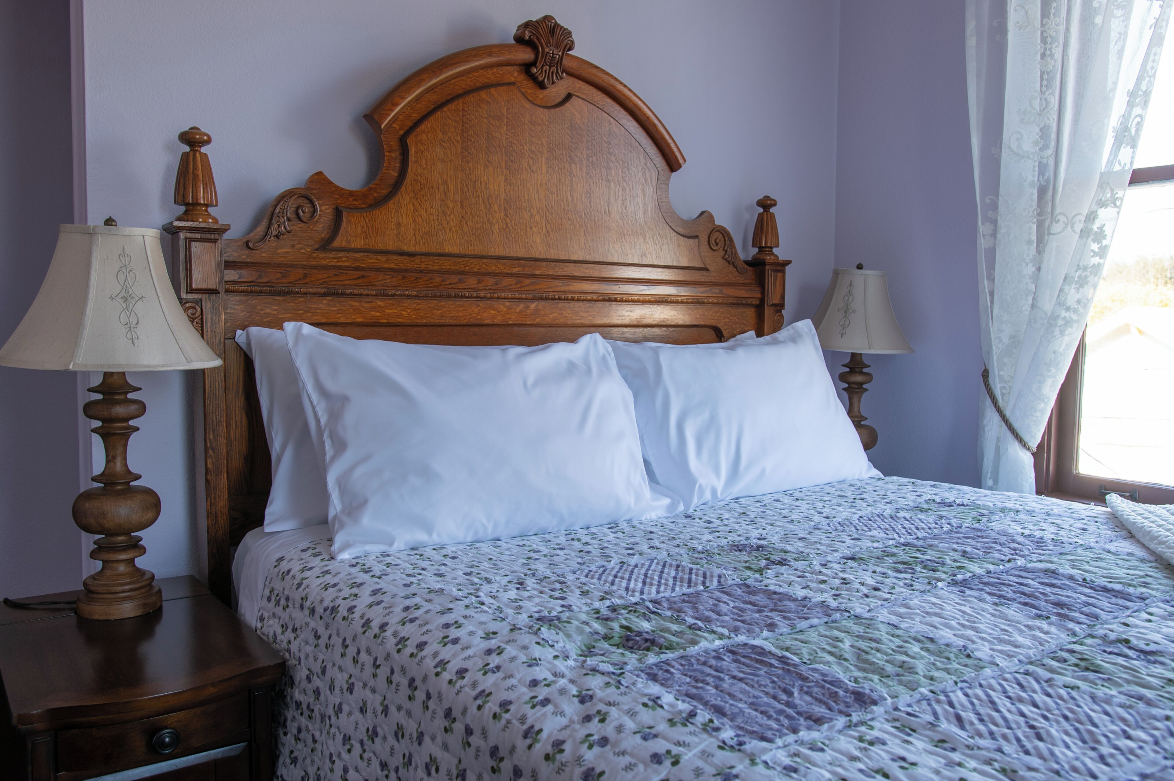 Een net opgemaakt bed met een quilt met bloemenmotief en twee witte kussens, met aan beide zijden houten nachtkastjes en lampen.