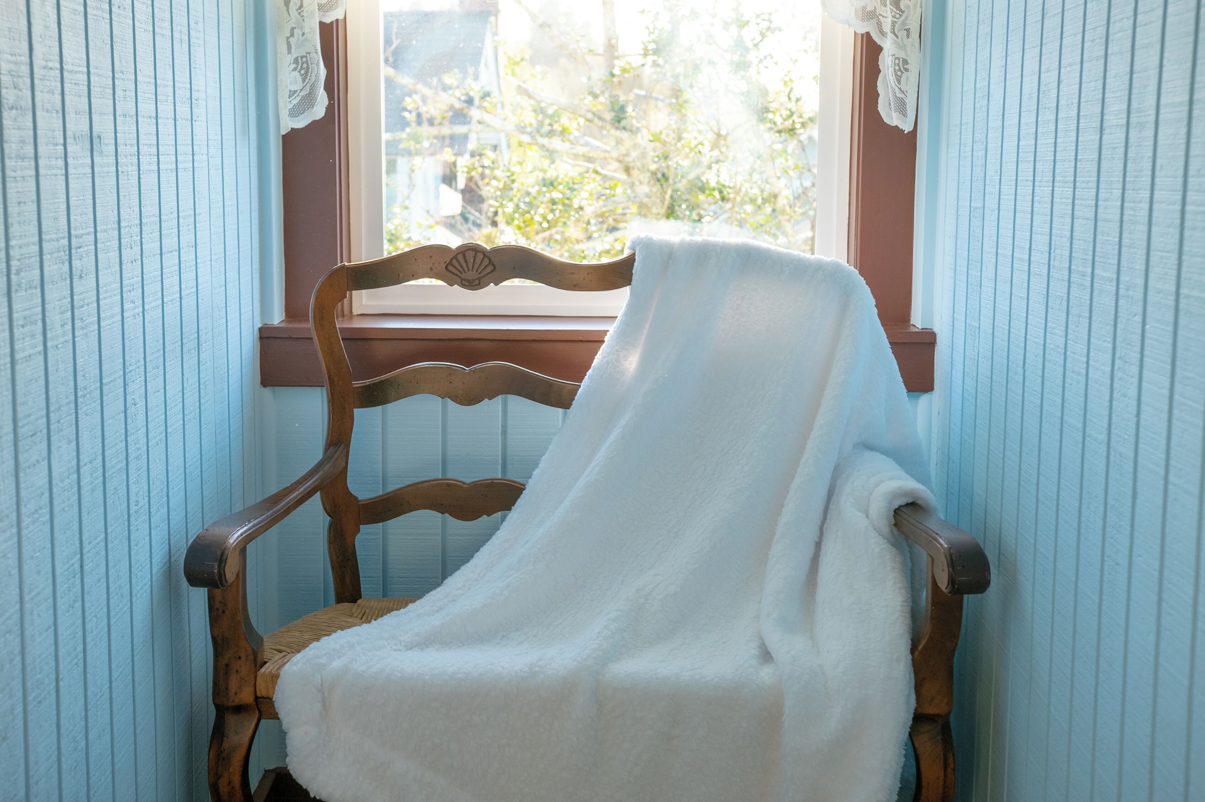 A wooden chair with a white towel draped over it, positioned by a window with lace curtains.
