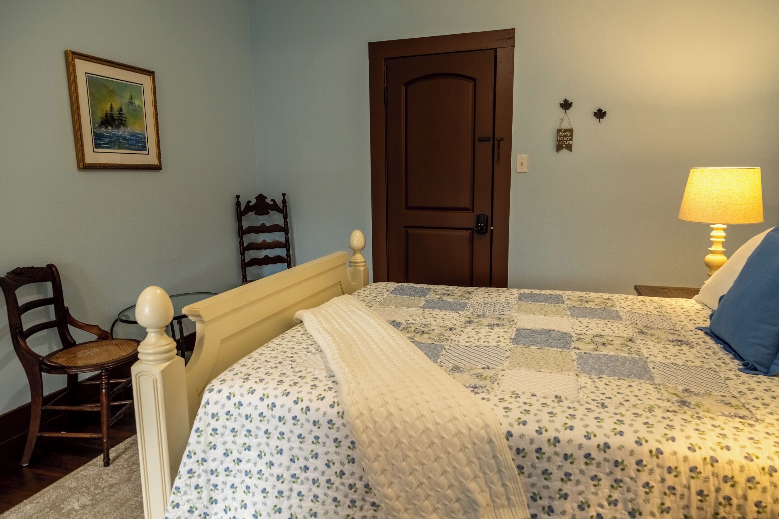 A bedroom with a bed, a chair, a door, a painting, and a lamp.