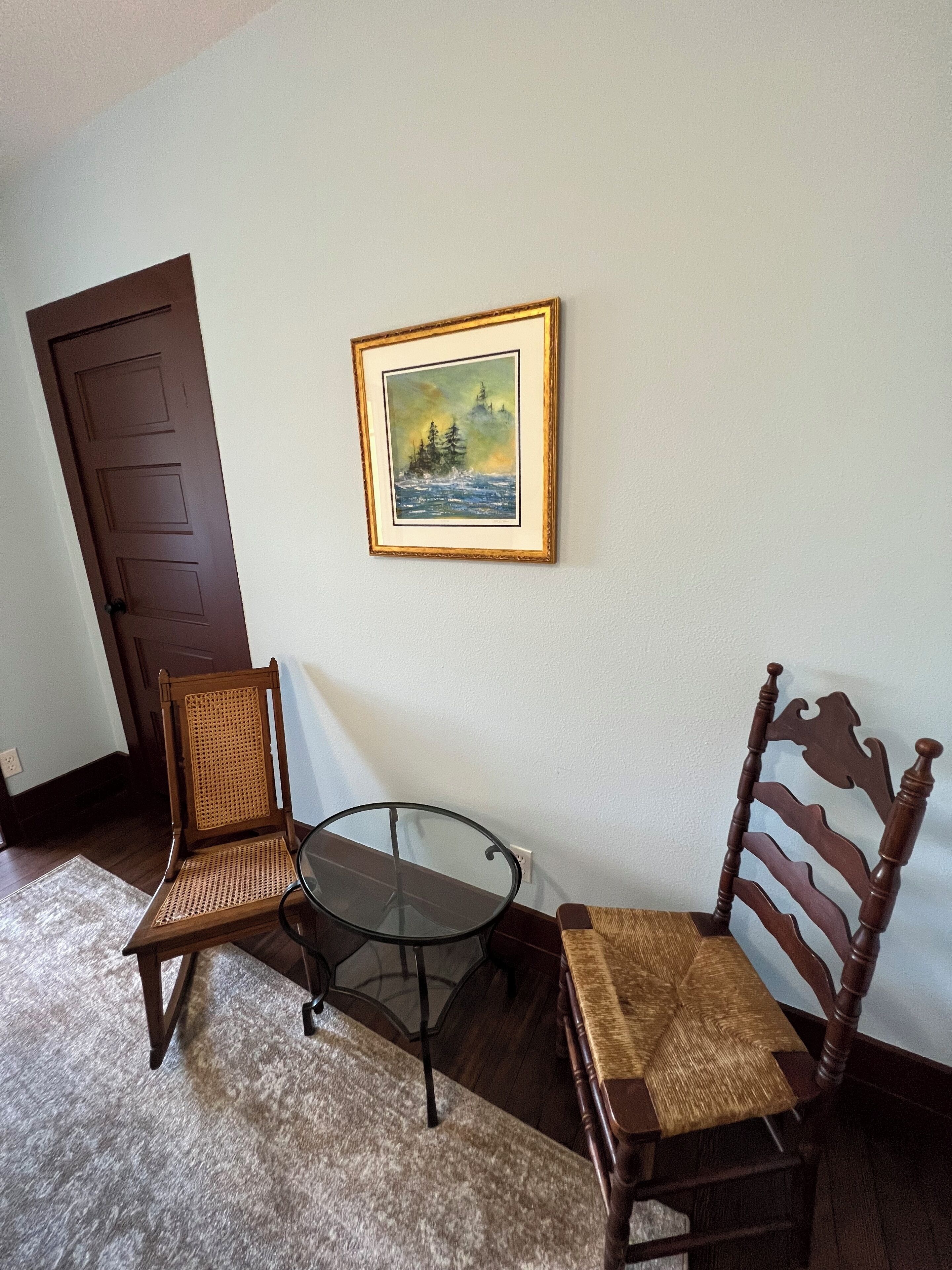 Une pièce avec une porte marron, un tableau encadré représentant un paysage, deux chaises en bois et une petite table en verre.