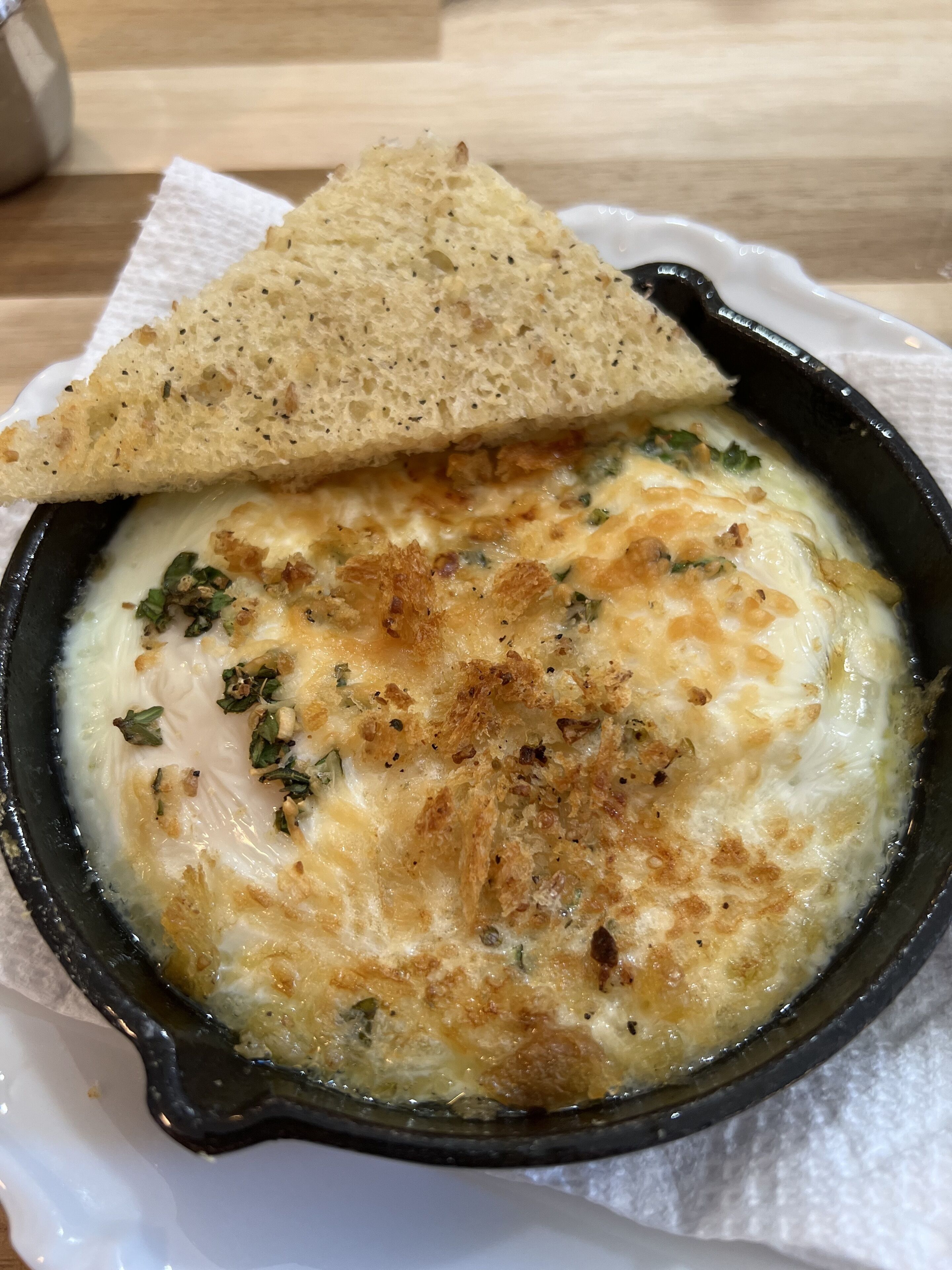 A dish of baked cheese with herbs and a slice of bread on a white plate.