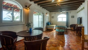 Amplia sala de estar con una mesa redonda de madera y sillas, un sofá y una televisión. La habitación tiene suelo de baldosas, vigas de madera en el techo y ventanas grandes con cortinas.