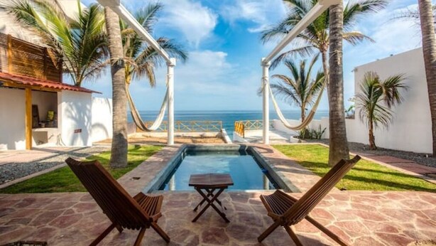 Zona de piscina con dos sillas de madera y una mesa, rodeada de palmeras y vista al océano.