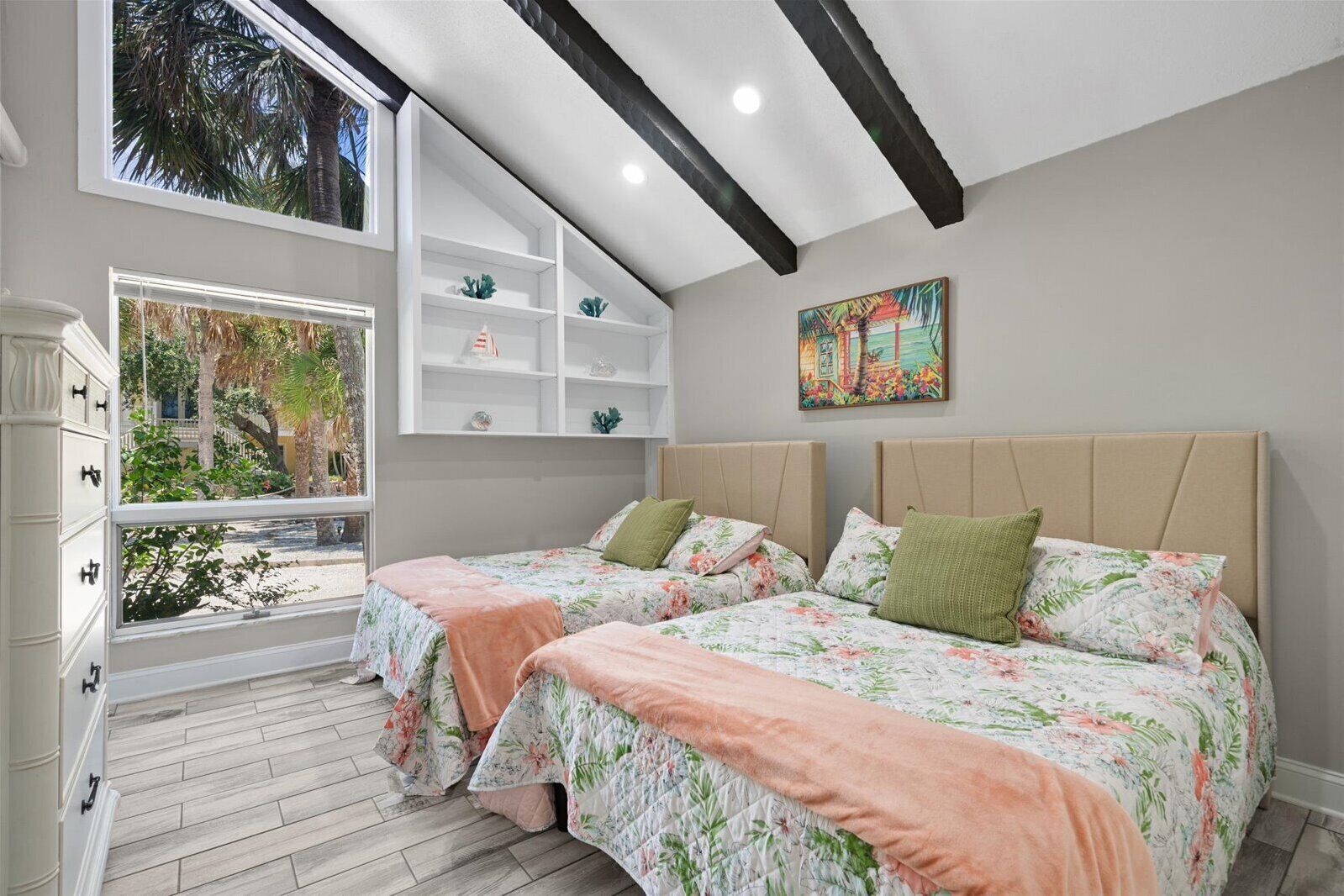 A bedroom with two beds, a window with a view of palm trees, a white dresser, and a shelf with decorative items.