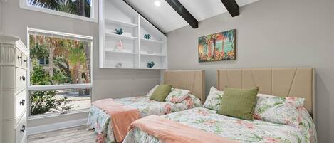 A bedroom with two beds, a window with a view of palm trees, a white dresser, and a shelf with decorative items.