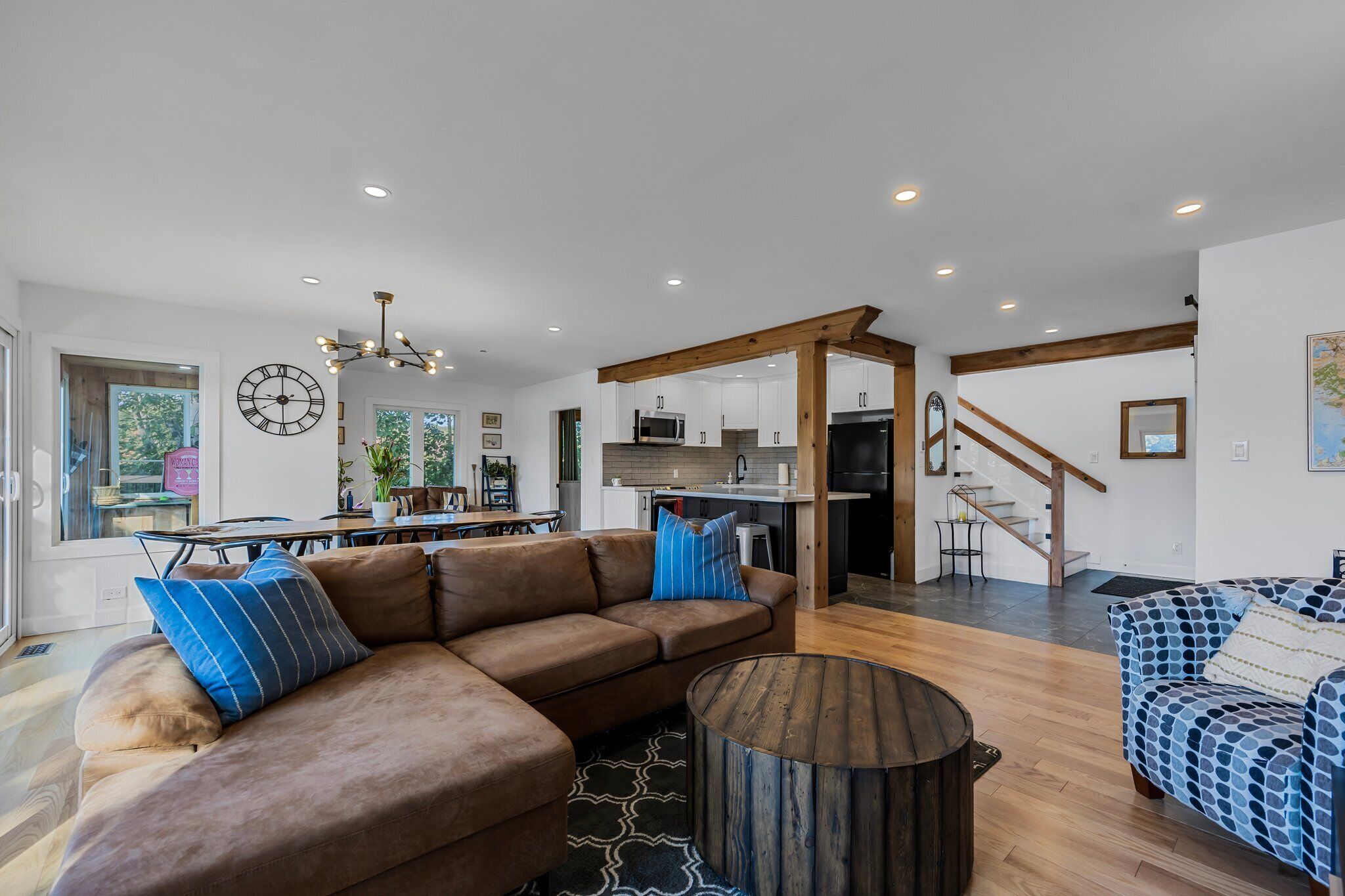 A spacious living room with a brown sectional sofa, a patterned blue sofa, and a wooden coffee table. A kitchen is visible in the background with modern appliances.