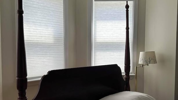 A four-poster bed with white linens, positioned between two windows with blinds, in a room with a white wall and a floor lamp. - Cannon Hill Inn (Rowlesburg)