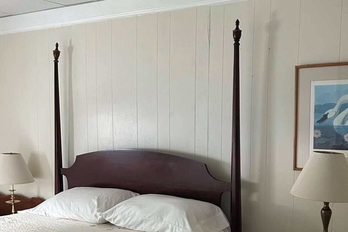 A four-poster bed with white linens, a wooden nightstand with a lamp, a framed picture on the wall, and a trash can.