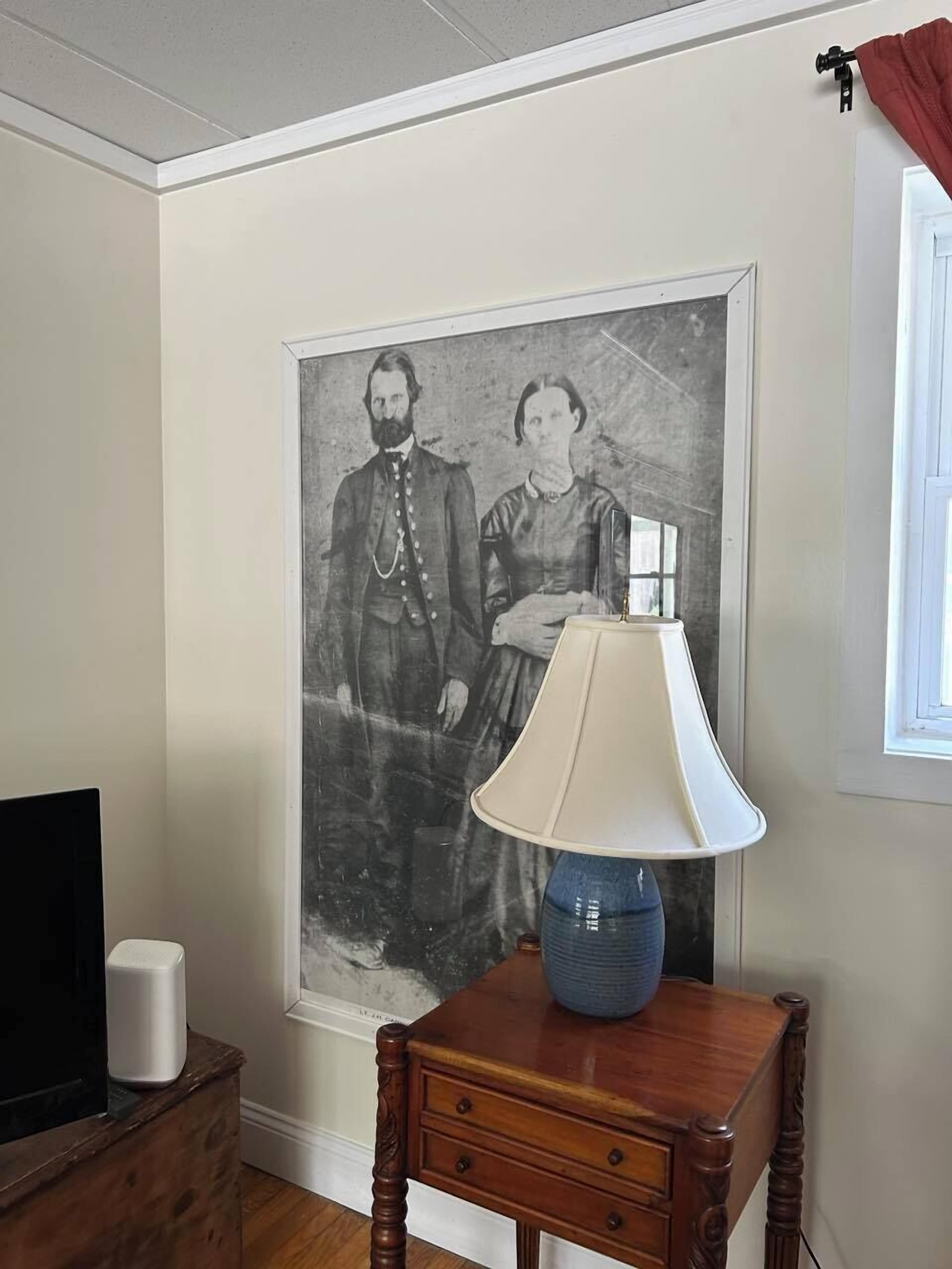 A room with a framed historical photograph on the wall, a wooden side table with a lamp, and a television on a stand.