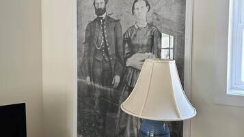 A room with a framed historical photograph on the wall, a wooden side table with a lamp, and a television on a stand.