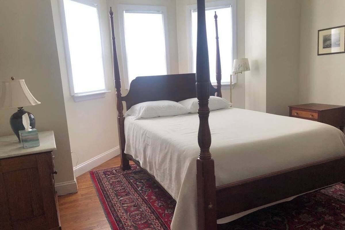 A four-poster bed with white linens, a wooden headboard, and a red patterned rug. There is a nightstand with a lamp and a small table with a vase.