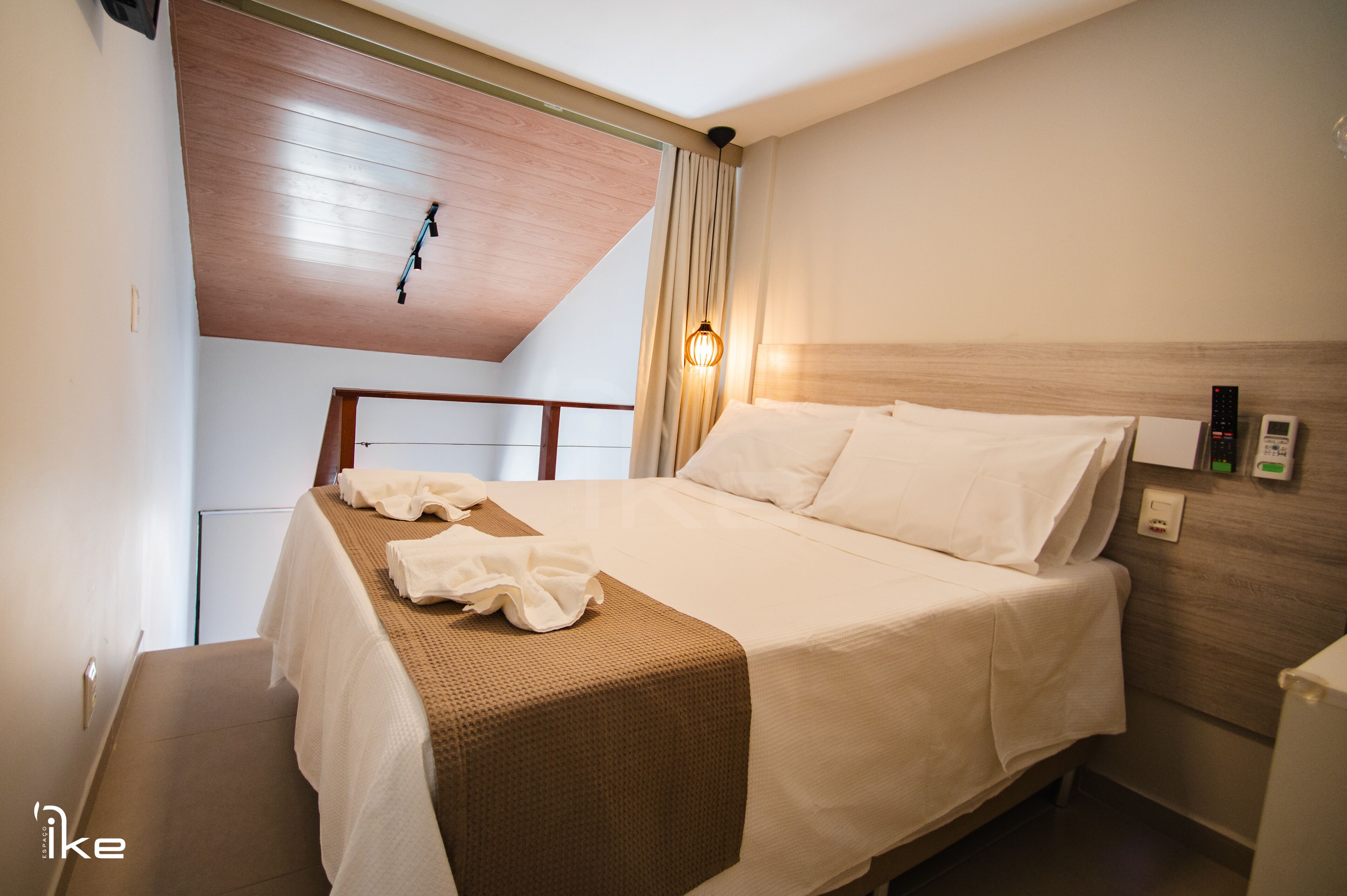 A hotel room with a bed, a wooden ceiling, a wall-mounted light, and a bedside table.