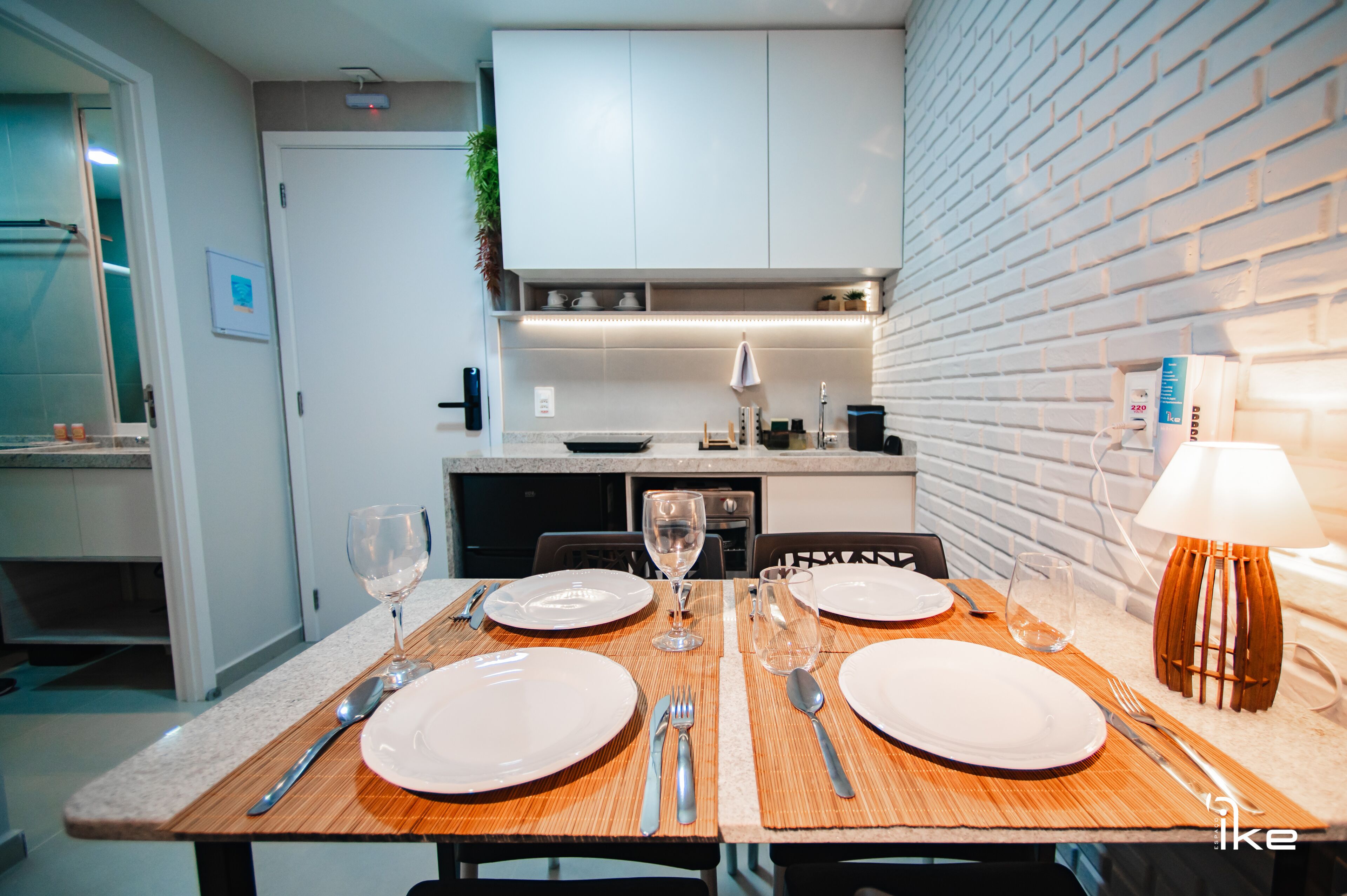 A dining area with a table set for two, a kitchen in the background, and a brick wall.