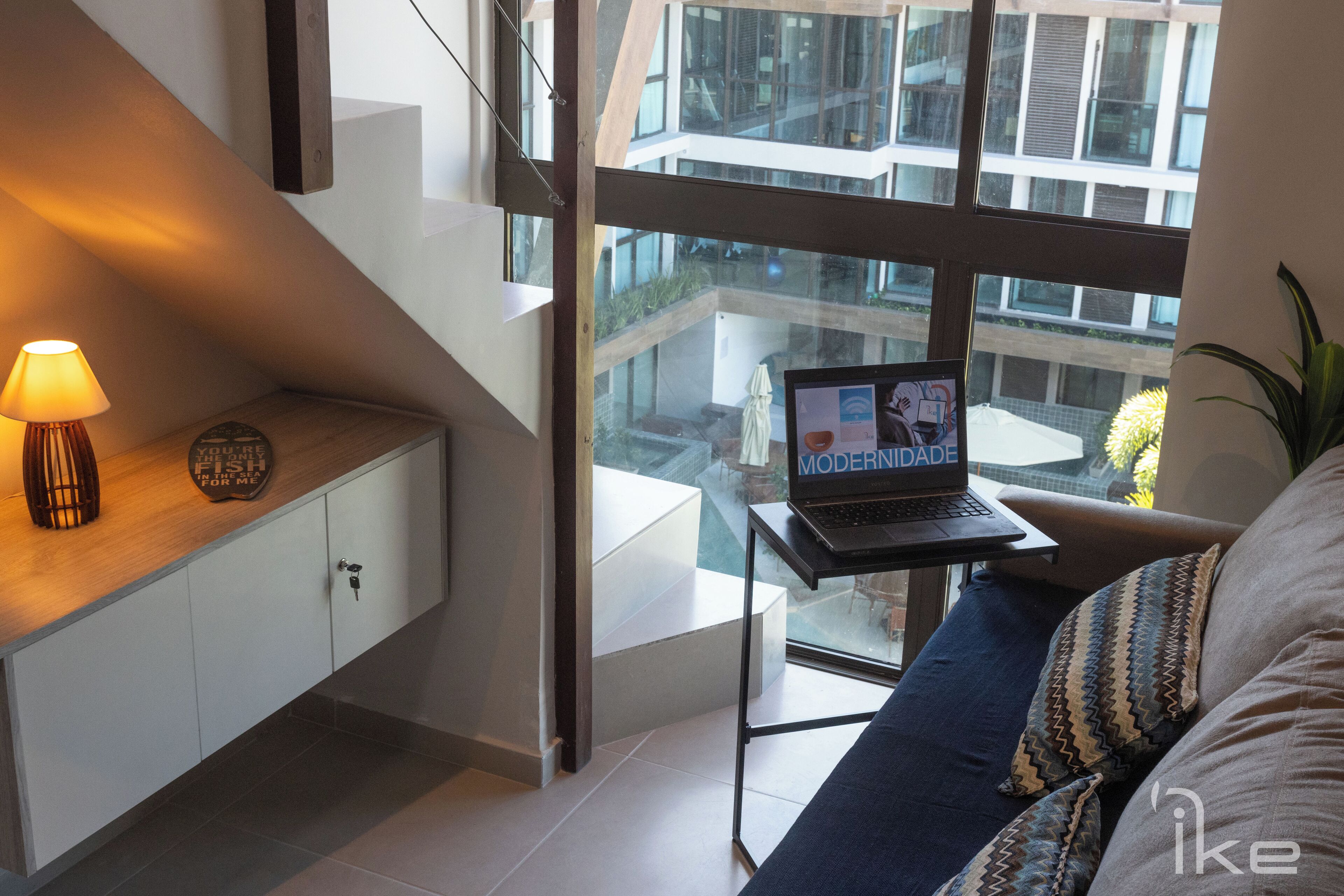 A modern living room with a laptop on a small table, a sofa with patterned cushions, a lamp on a wooden console, and a view of a cityscape through large windows.