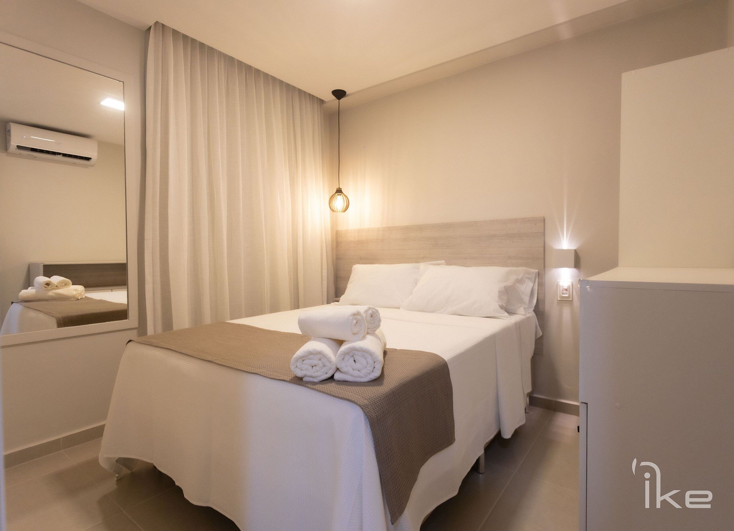 A hotel room with a neatly made bed, a bedside table, a mirror, an air conditioning unit, and a wall-mounted light fixture.