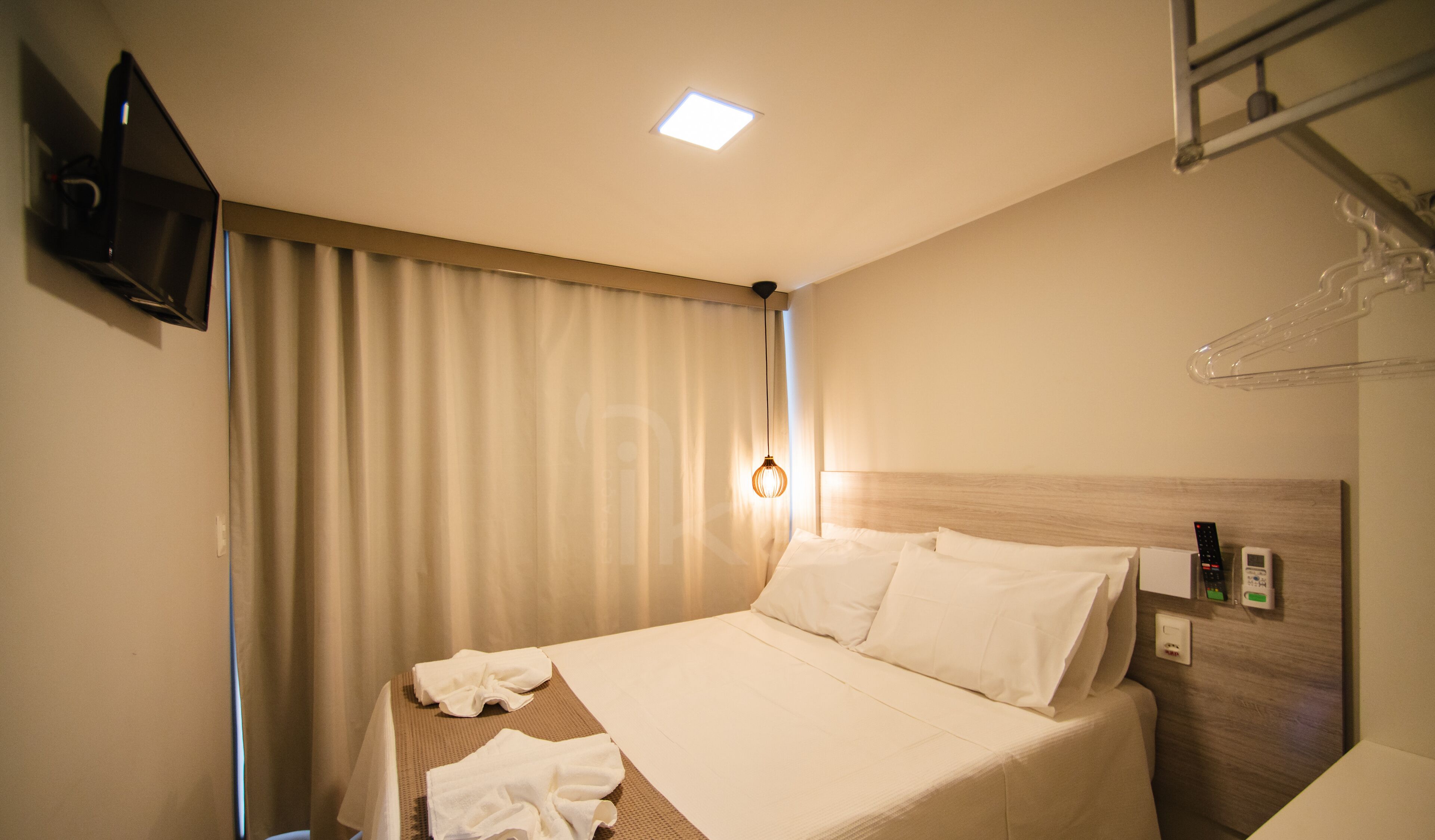 A hotel room with a bed, a TV mounted on the wall, a ceiling light, and a towel rack.
