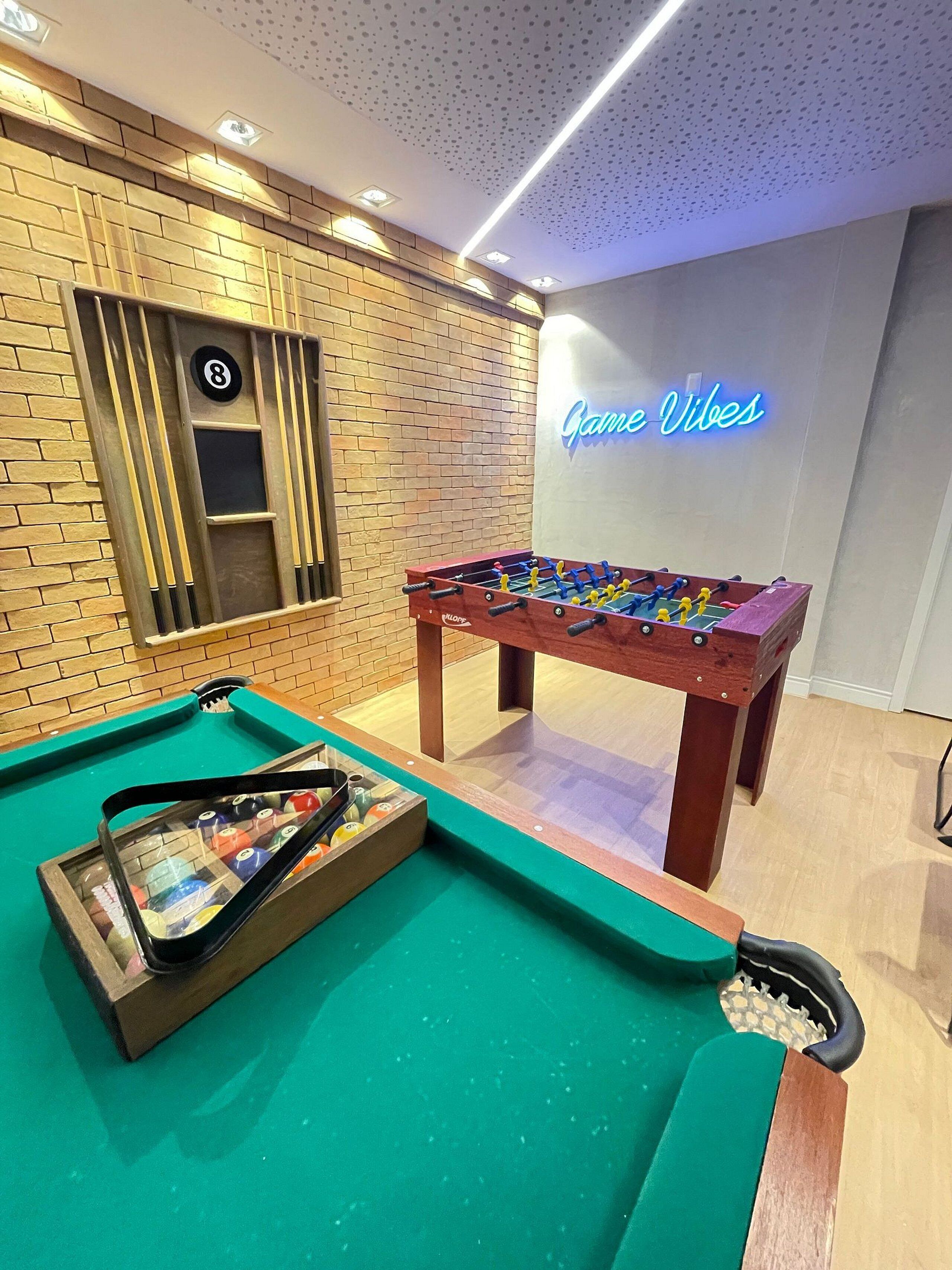 A foosball table, pool table, and a neon sign reading "Game Vibes" in a room with brick walls and recessed lighting.