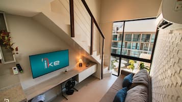 A modern living room with a flat-screen TV, a staircase, and a large window offering a view of the outdoors.
