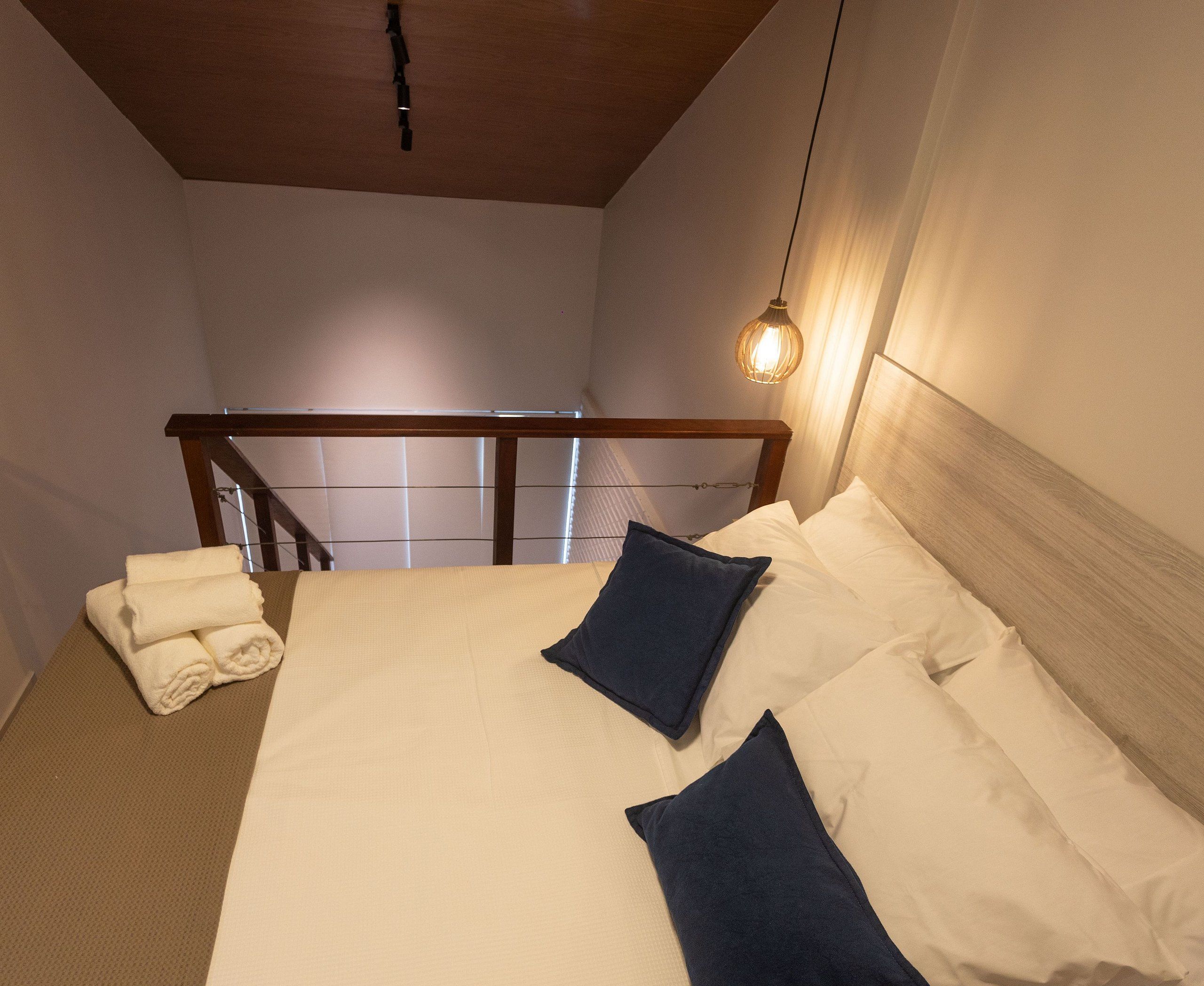 Un lit bien fait, avec des oreillers blancs et bleu foncé, une tête de lit en bois et un luminaire suspendu.