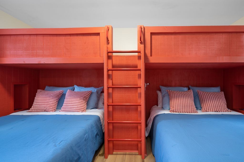 A room with two bunk beds, each with a blue bedspread and red checkered pillows. A wooden ladder connects the beds.