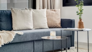 A modern living room with a grey sofa, beige and white cushions, a small round table with a glass and book, and a vase with green plants. - Sanders Premier - Lovely 2 bdr apt With Balcony (Limassol)