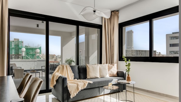 A modern living room with a grey sofa, a coffee table, and a view of the cityscape through large windows. - Sanders Premier - Lovely 2 bdr apt With Balcony (Limassol)