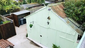 Pieni, suljettu ulkotila, jossa on betonilattia, puinen penkki ja aaltopeltikattoinen rakennus, jonka sisällä on roikkuvia kasveja.